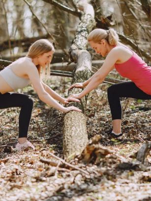 Side view of young female athletes in activewear stretching legs together using fallen tree during fitness workout in spring forest adhering to healthy lifestyle