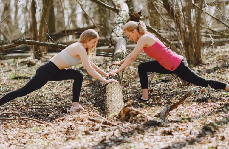 Side view of young female athletes in activewear stretching legs together using fallen tree during fitness workout in spring forest adhering to healthy lifestyle